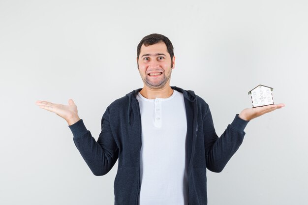 Jeune homme tenant le modèle de la maison dans une main et étirant une autre main vers la gauche en t-shirt blanc et sweat à capuche noir zippé à l'avant et à l'optimiste, vue de face.