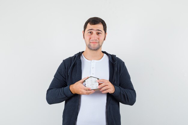 Jeune homme tenant le modèle de maison avec les deux mains en t-shirt blanc et sweat à capuche noir zippé sur le devant et à la vue sérieuse, avant.