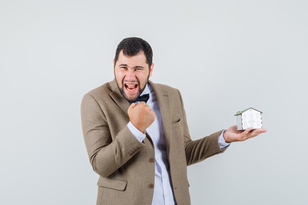 Jeune homme tenant le modèle de maison avec le geste gagnant en costume et à la vue de face, heureux.