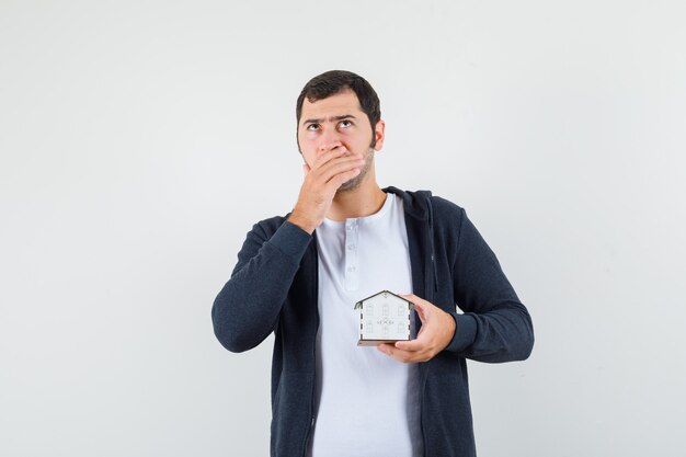 Jeune homme tenant un modèle de maison, pensant à quelque chose en t-shirt blanc et sweat à capuche noir zippé sur le devant et à la pensif. vue de face.