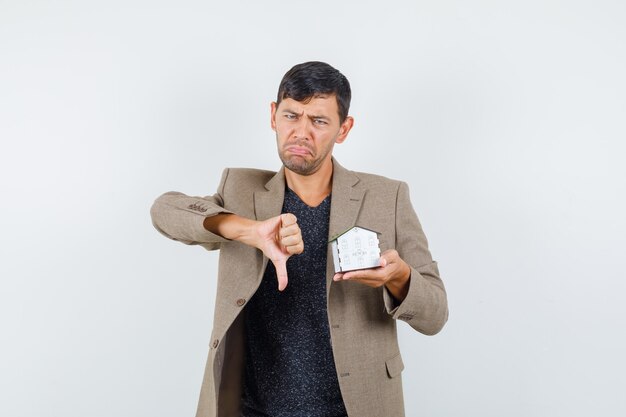 Jeune homme tenant un modèle de maison avec le pouce vers le bas en veste, t-shirt et à la vue mécontent, de face.