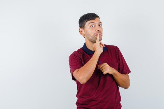 Jeune homme tenant le petit doigt sur les lèvres en t-shirt et l'air curieux, vue de face.