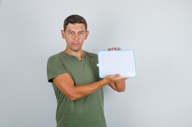 Jeune homme tenant un petit tableau blanc en t-shirt vert armée, vue de face.