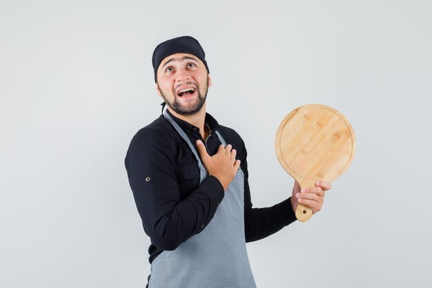 Jeune homme tenant une planche à découper en chemise, tablier et à la recherche de rêve. vue de face.