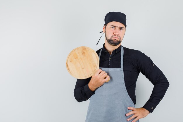 Jeune homme tenant une planche à découper en chemise, tablier et regardant stricte, vue de face.