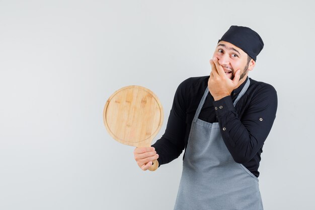 Jeune homme tenant une planche à découper avec la main sur la bouche en chemise, tablier et air heureux. vue de face.