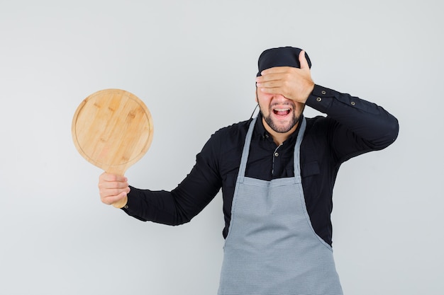 Jeune homme tenant une planche à découper avec la main sur les yeux en chemise, tablier et air heureux. vue de face.