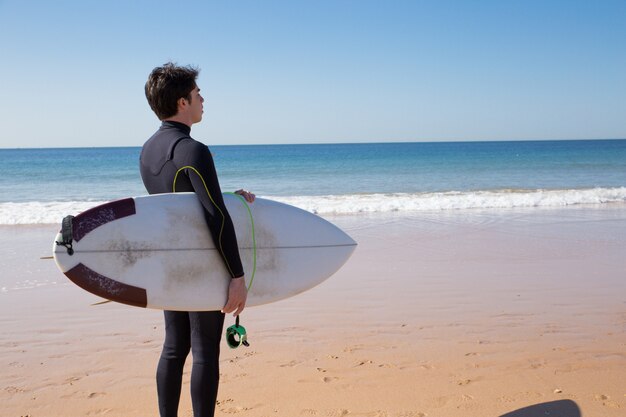 Jeune homme tenant une planche de surf et regardant la mer