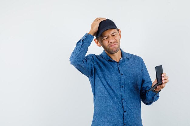 Jeune homme tenant un portefeuille en chemise bleue, casquette et oublieux, vue de face.