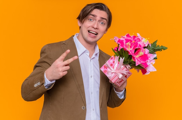 Jeune homme tenant présent et bouquet de fleurs à l'avant montrant v-sign souriant joyeusement va féliciter avec la journée internationale de la femme debout sur le mur orange