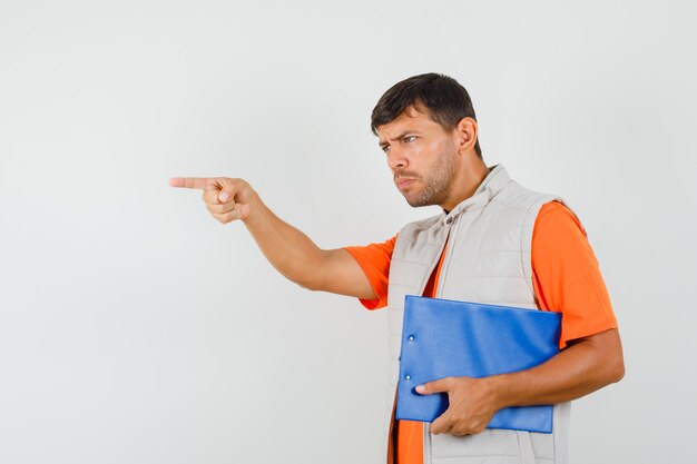 Jeune homme tenant le presse-papiers, donnant des instructions en t-shirt, veste et à la recherche concentrée. vue de face.