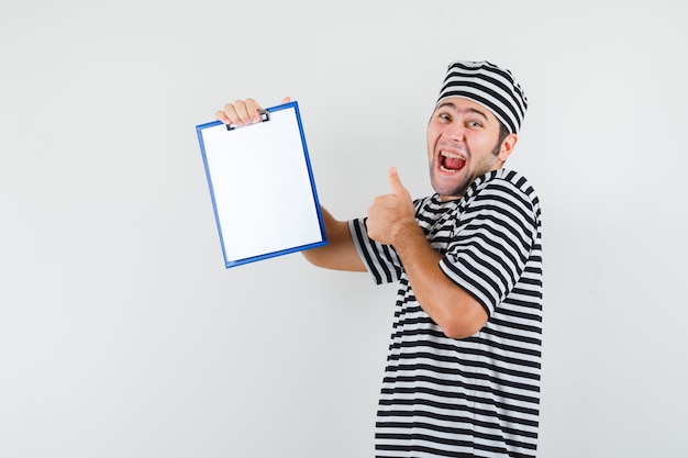 Jeune homme tenant le presse-papiers, montrant le pouce vers le haut en t-shirt, chapeau et à la joyeuse, vue de face.