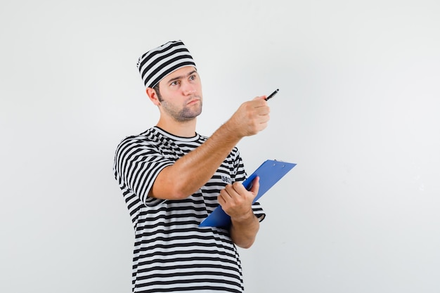 Jeune homme tenant le presse-papiers, stylo, pointant vers l'extérieur en t-shirt, chapeau et regardant pensif, vue de face.