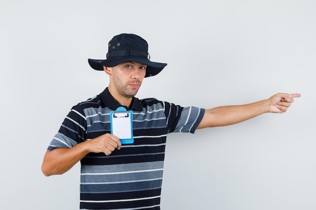 Jeune homme tenant le presse-papiers tout en pointant sur le côté en t-shirt, vue de face de chapeau.