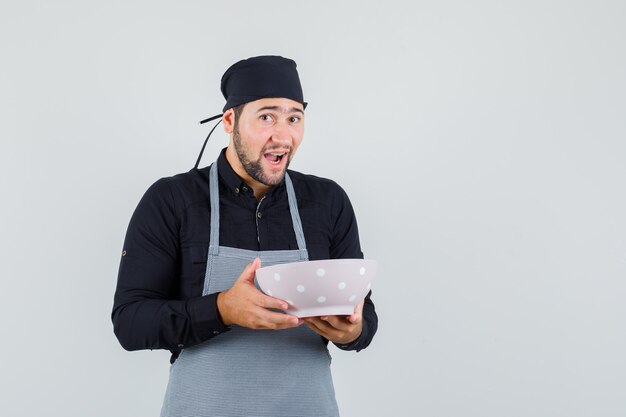 Jeune homme tenant le repas dans un bol en chemise, tablier et à la joyeuse, vue de face.