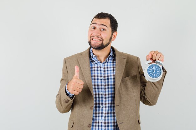 Jeune homme tenant un réveil, montrant le pouce vers le haut en chemise, veste et à la vue de face, heureux.