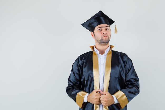 Photo gratuite jeune homme tenant sa robe en uniforme d'études supérieures et à la confiance. vue de face.