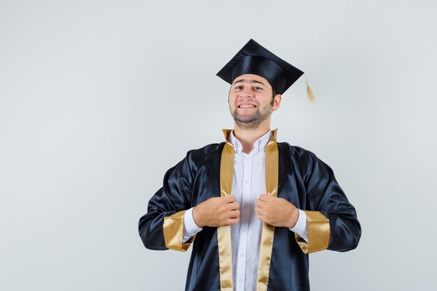 Jeune homme tenant sa robe en uniforme d'études supérieures et à la fierté, vue de face.