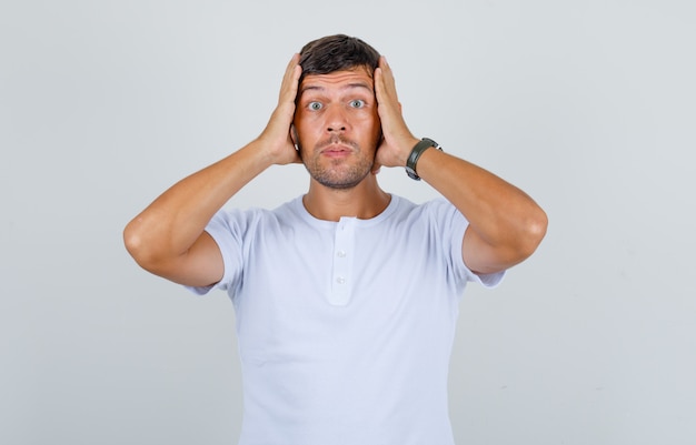 Jeune homme tenant sa tête sur les mains en t-shirt blanc et regardant anxieux, vue de face.