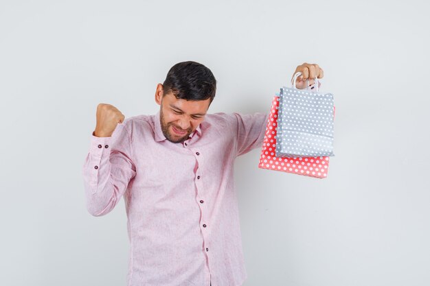 Jeune homme tenant des sacs en papier avec le geste gagnant en chemise et à la vue de face, heureux.