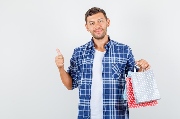 Jeune homme tenant des sacs en papier avec le pouce en haut en chemise et à la vue de face, heureux.