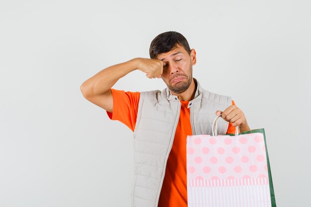 Jeune homme tenant des sacs à provisions, se frottant les yeux en t-shirt, veste et à la tristesse. vue de face.