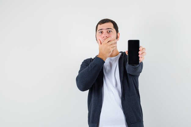 Jeune homme tenant un smartphone, couvrant la bouche avec la main en t-shirt blanc et sweat à capuche noir à glissière sur le devant et à la surprise. vue de face.