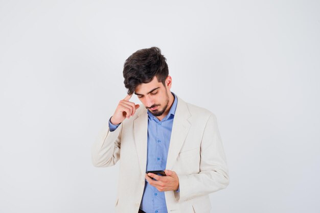 Jeune homme tenant un smartphone et mettant l'index sur l'oreille en t-shirt bleu et veste de costume blanc et ayant l'air sérieux