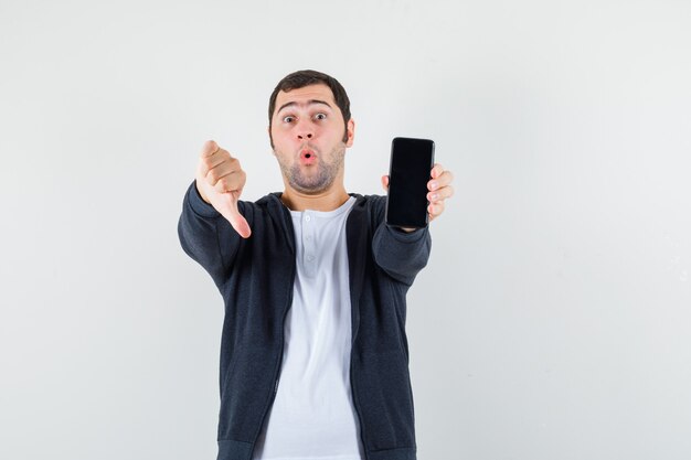 Jeune homme tenant un smartphone, montrant le pouce vers le bas en t-shirt blanc et sweat à capuche noir à glissière sur le devant et à la surprise. vue de face.