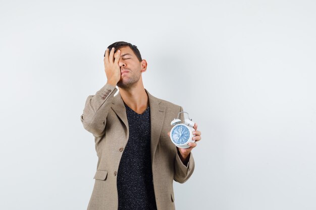 Jeune homme tenant son visage tout en montrant l'horloge en veste marron grisâtre et à la recherche d'anxiété. vue de face.