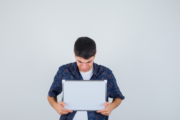 Jeune homme tenant un tableau blanc, le regardant en t-shirt blanc et chemise à fleurs