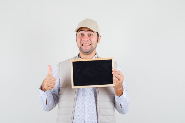 Jeune homme tenant le tableau et montrant le pouce vers le haut en veste beige et casquette et regardant heureux, vue de face.