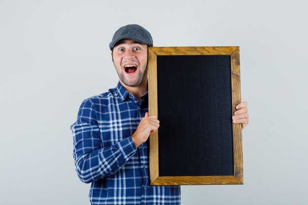 Jeune homme tenant le tableau noir en chemise, casquette et à la joyeuse, vue de face.