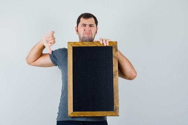 Jeune homme tenant un tableau noir montrant le pouce vers le bas en t-shirt gris et à la recherche de mécontentement