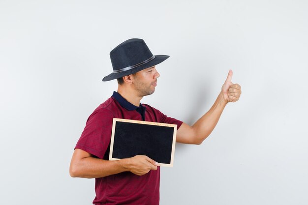 Jeune homme tenant un tableau noir avec le pouce vers le haut en t-shirt, chapeau, vue de face.