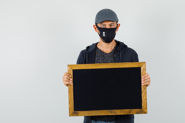 Jeune homme tenant un tableau noir en t-shirt, veste, casquette, masque, vue de face.