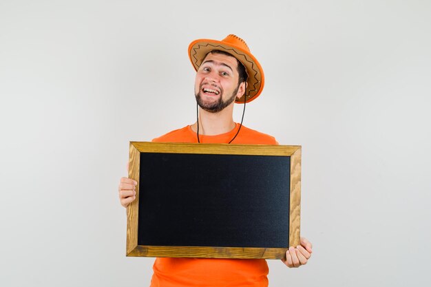 Jeune homme tenant un tableau en t-shirt orange, un chapeau et l'air gai. vue de face.