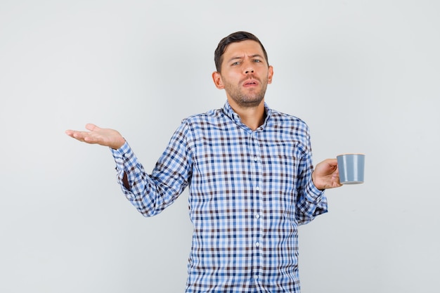 Jeune homme tenant une tasse de boisson en geste perplexe en chemise à carreaux