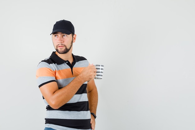 Jeune homme tenant une tasse de boisson tout en regardant de côté en t-shirt, casquette et à la recherche concentrée. vue de face.