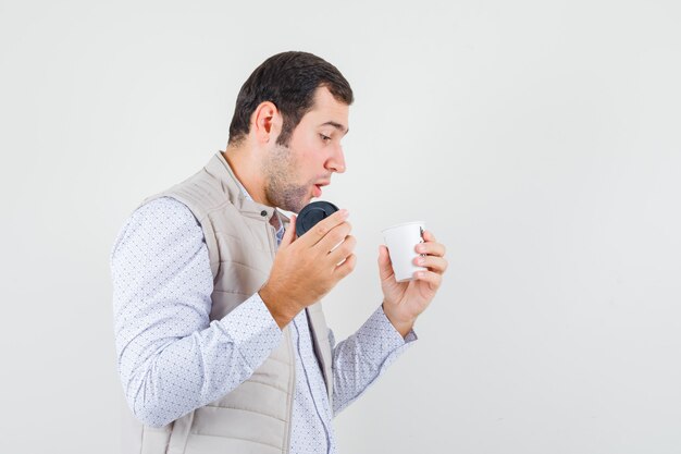 Jeune homme tenant une tasse de café à emporter et essayant de l'ouvrir en veste beige et à la recherche concentrée. vue de face.