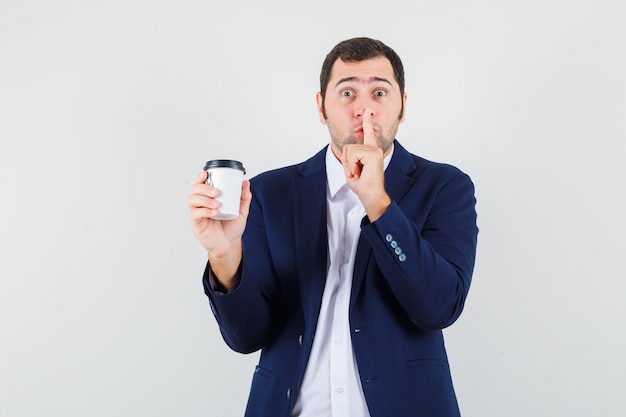 Jeune homme tenant une tasse de café, montrant le geste de silence en chemise, vue de face de la veste