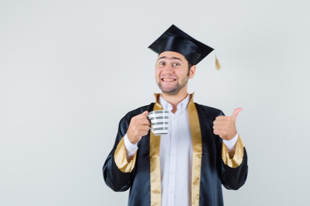 Jeune homme tenant une tasse de café, montrant le pouce vers le haut en uniforme d'études supérieures et regardant joyeux, vue de face.