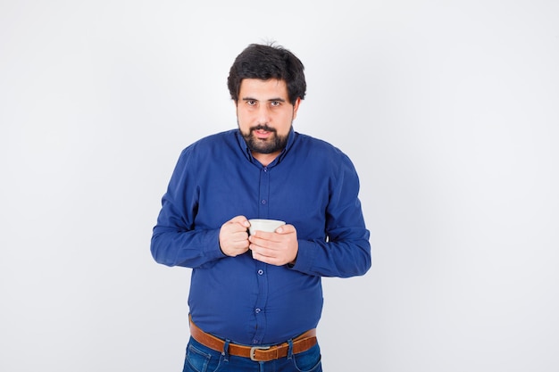 Jeune homme tenant une tasse à deux mains en chemise bleue et jeans et à la recherche de sérieux. vue de face.