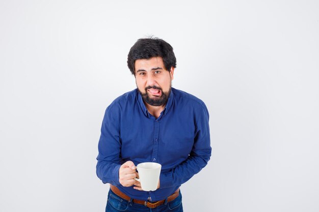 Jeune homme tenant une tasse et mettant la main sur le ventre en chemise bleue et jeans et l'air épuisé, vue de face.