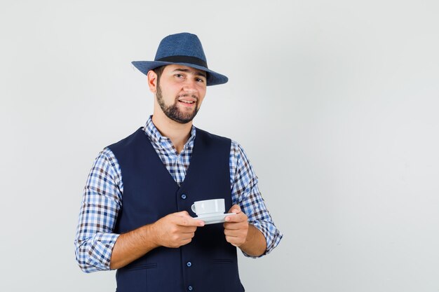 Jeune homme tenant une tasse avec soucoupe en chemise, gilet, chapeau et à la satisfaction. vue de face.