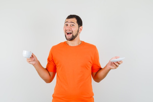 Jeune homme tenant une tasse et une soucoupe en t-shirt orange et regardant joyeux, vue de face.