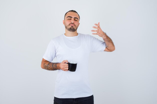 Jeune homme tenant une tasse de thé et étirant la main comme tenant quelque chose d'imaginaire tout en fermant les yeux en t-shirt blanc et pantalon noir et l'air calme