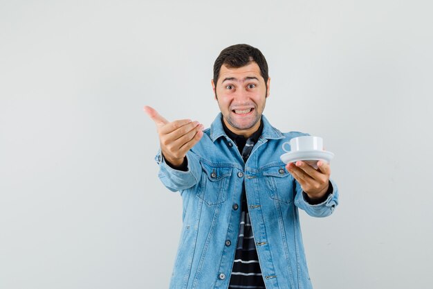 Jeune homme tenant une tasse de thé, invitant à venir en t-shirt, veste et à joyeux