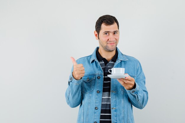 Jeune homme tenant une tasse de thé, montrant le pouce vers le haut en t-shirt, veste et à la satisfaction