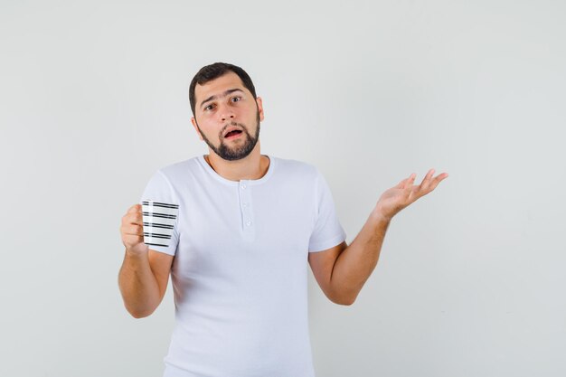 Jeune homme tenant une tasse tout en montrant un geste impuissant en t-shirt blanc et l'air confus, vue de face.